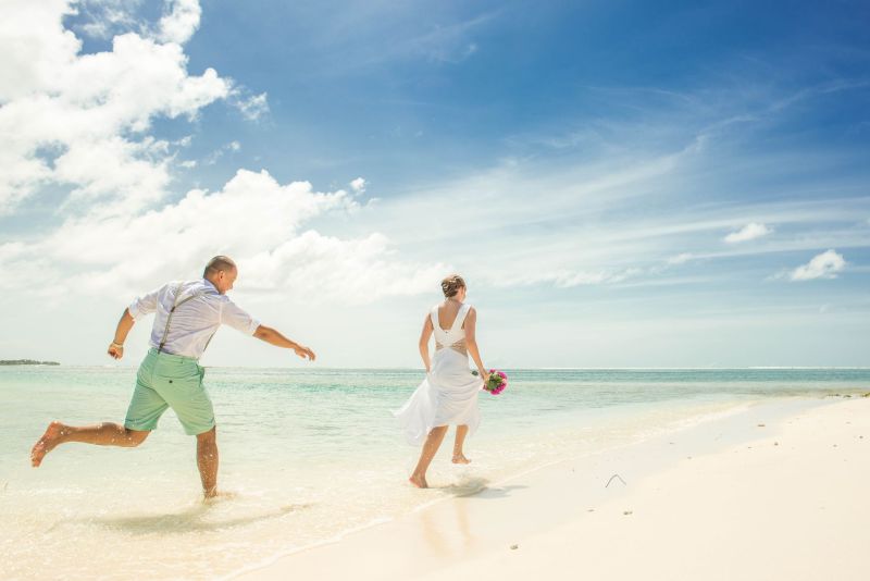 Example of beach casual attire: Bride and groom running on beach at beach wedding, groom in green pastel shorts, bride in short white dress